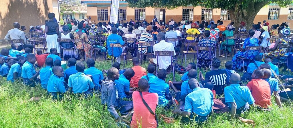 Staff and children are seated outside under a tree listening to a health talk at a primary school in Agago District, Uganda