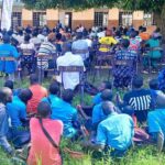 Staff and children are seated outside under a tree listening to a health talk at a primary school in Agago District, Uganda