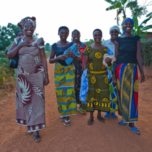 Seven rwandan women in bright dressed clothes walk down a dusty red road - all are smiling. (c) Andrew Sutton