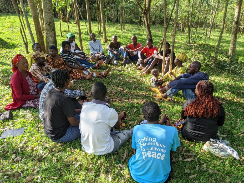 A group of people sitting in a circle outside on grass, among trees